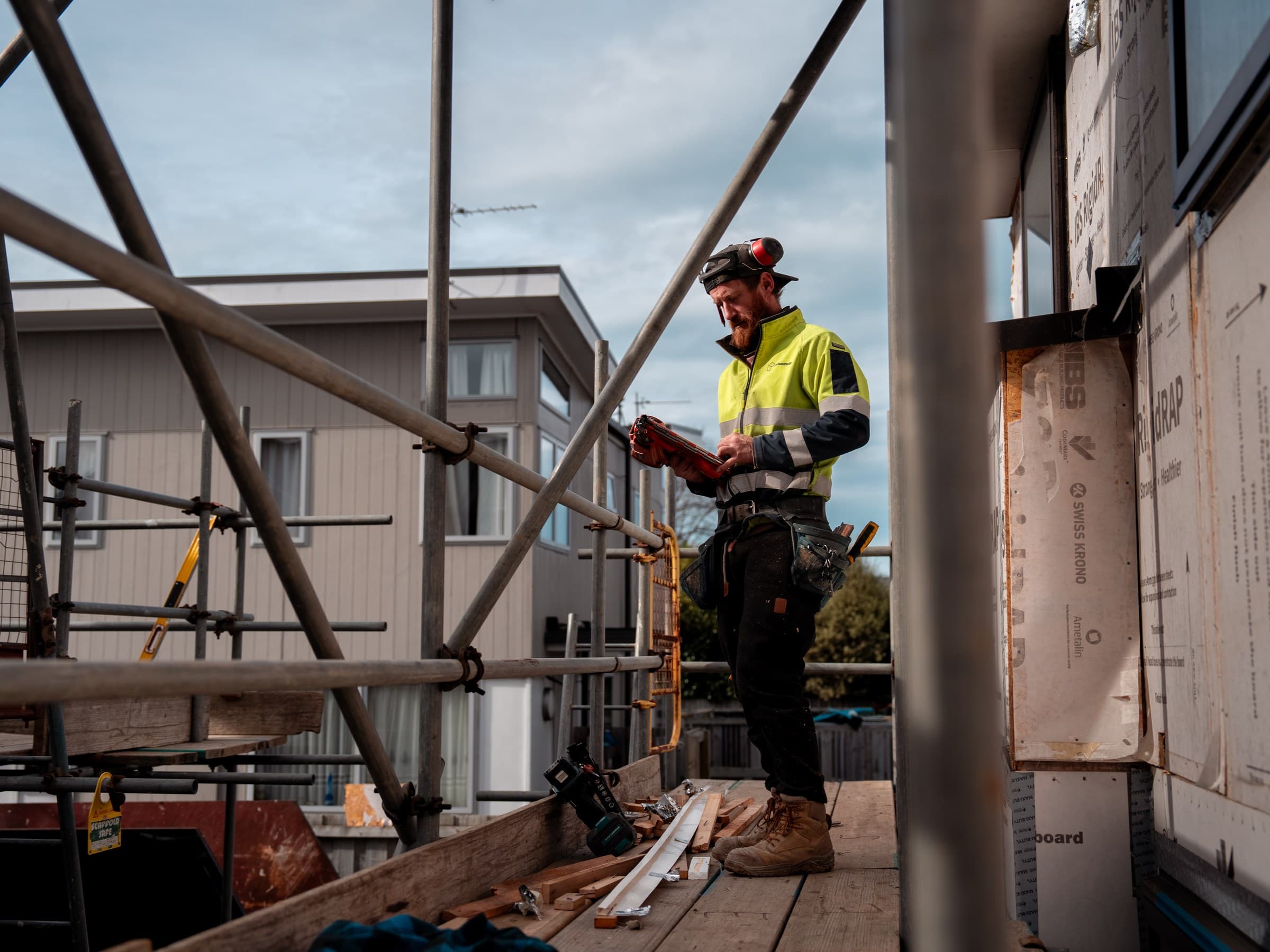 A worker in hi-vis standing on a scaffold platform beside a timber-framed wall sheathed in building wrap, looking down at a small tool held in both hands, scaffold tubes crossing the frame, and a neighbouring two-storey house behind under an overcast sky