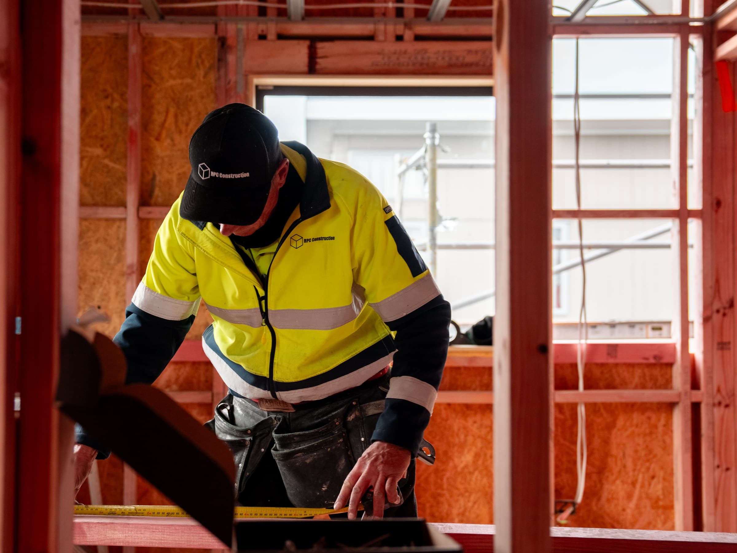 A worker in an RPC Construction hi-vis jacket and branded cap, head down, marking a line with a steel tape on a timber bench inside a building under construction, exposed timber framing and OSB sheathing all around, and a window opening behind looking out to scaffolding and a neighbouring building