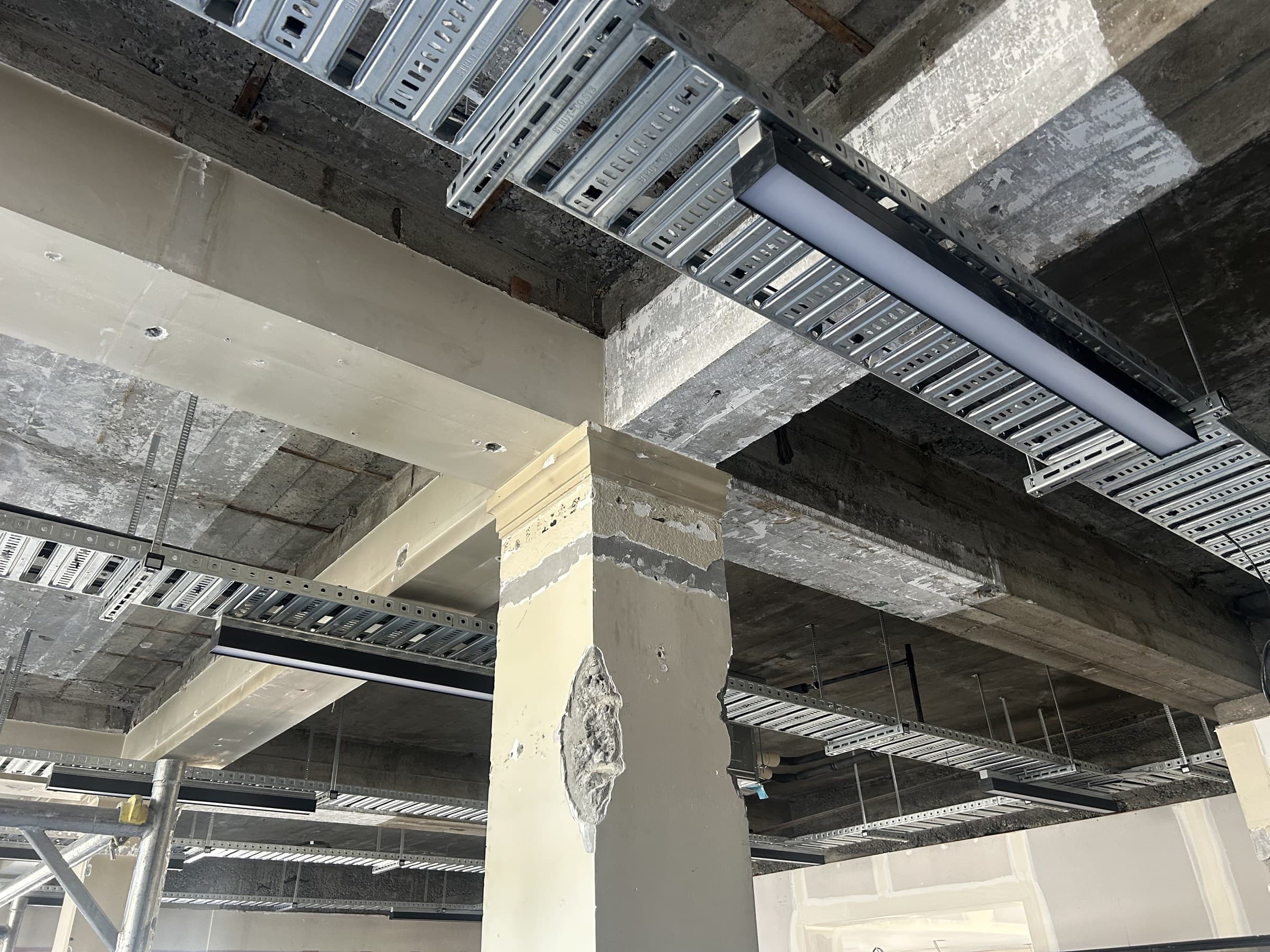Exposed concrete beams and a column inside a building under fit-out, with galvanised cable tray and a suspended linear light fitting running between them