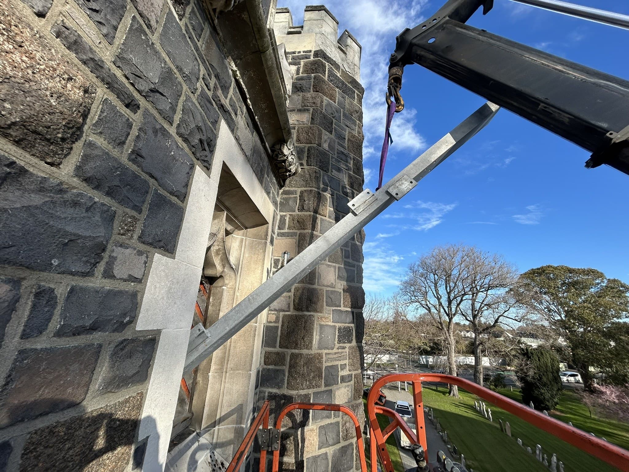 A galvanised steel member on a crane sling being brought up the stone tower of an old church, worked on from a boom lift, with the churchyard and street below