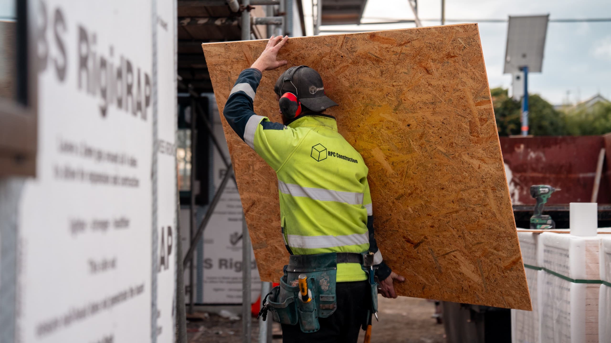 A worker in RPC Construction hi-vis carrying a large sheet of OSB past a scaffold, stacked framing timber and wrapped building board along the site behind him