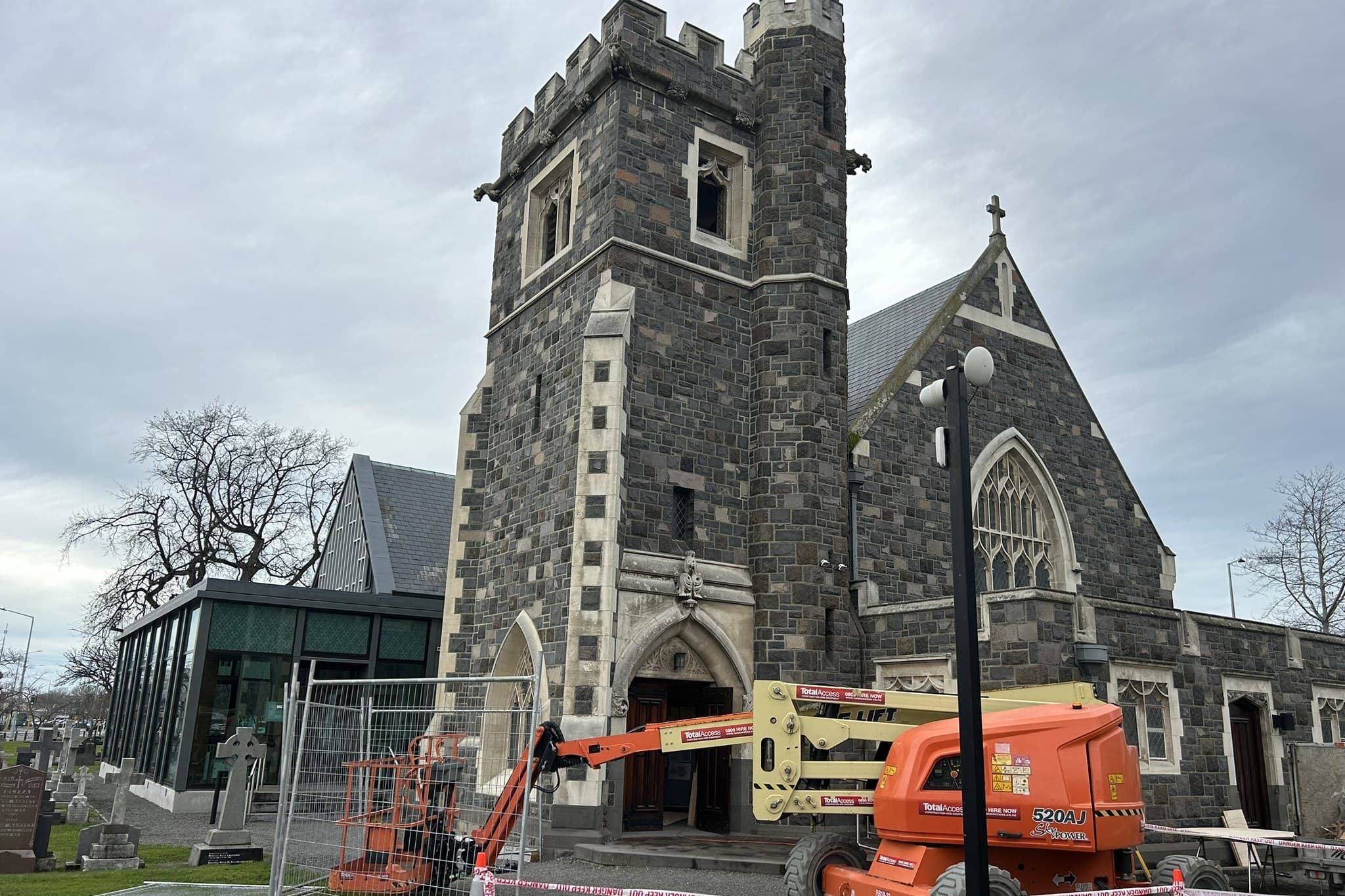 A stone church with its front doors standing open and boards laid over the threshold, a boom lift set up at the entrance, and the working area in front of it closed off with temporary fence panels, hazard tape and traffic cones