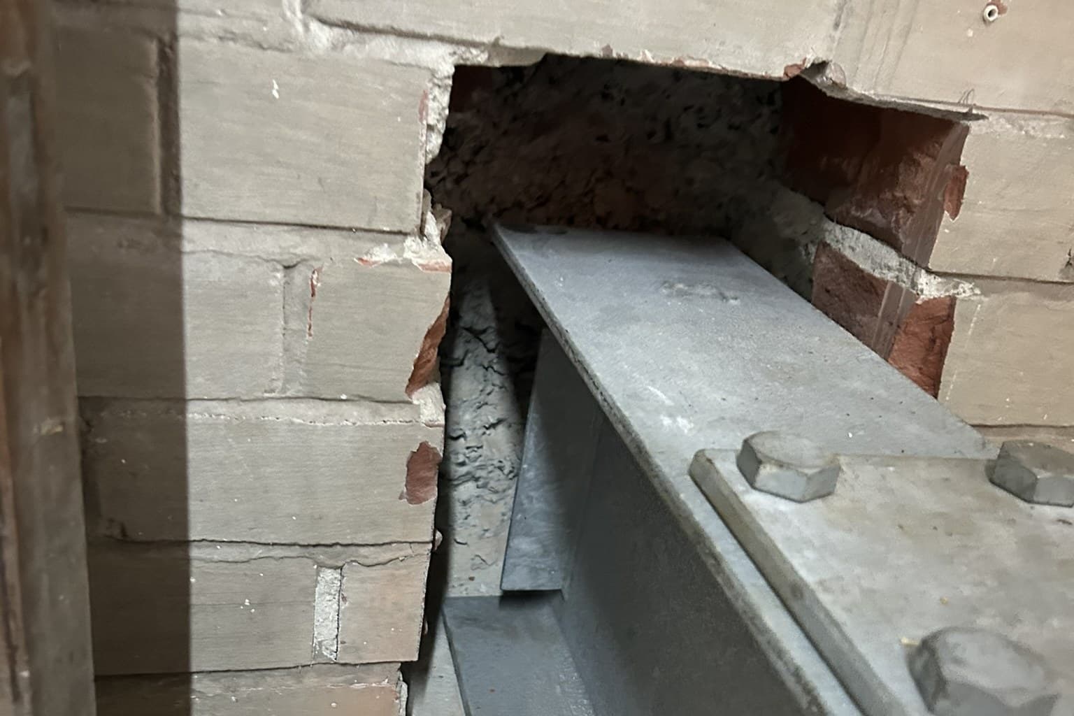 A new galvanised steel beam bearing into a pocket cut through an old brick wall, the cut face of the brickwork open with its red core showing, and a bolted top plate with hex bolts in the near foreground
