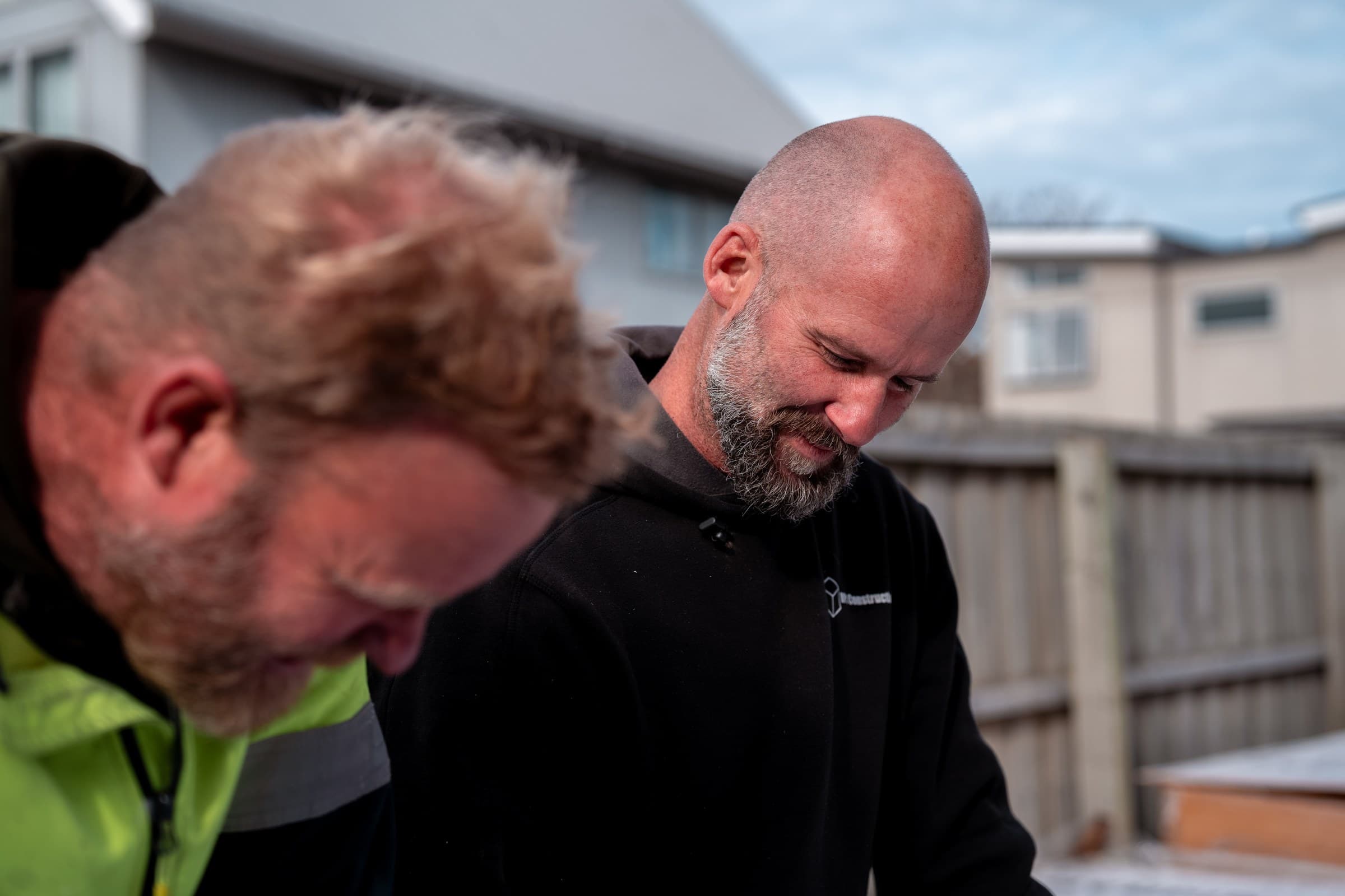 Two workers close together on a site, one in an RPC Construction hoodie looking down at the work in front of him, the other in hi-vis in the near foreground, houses and a paling fence behind