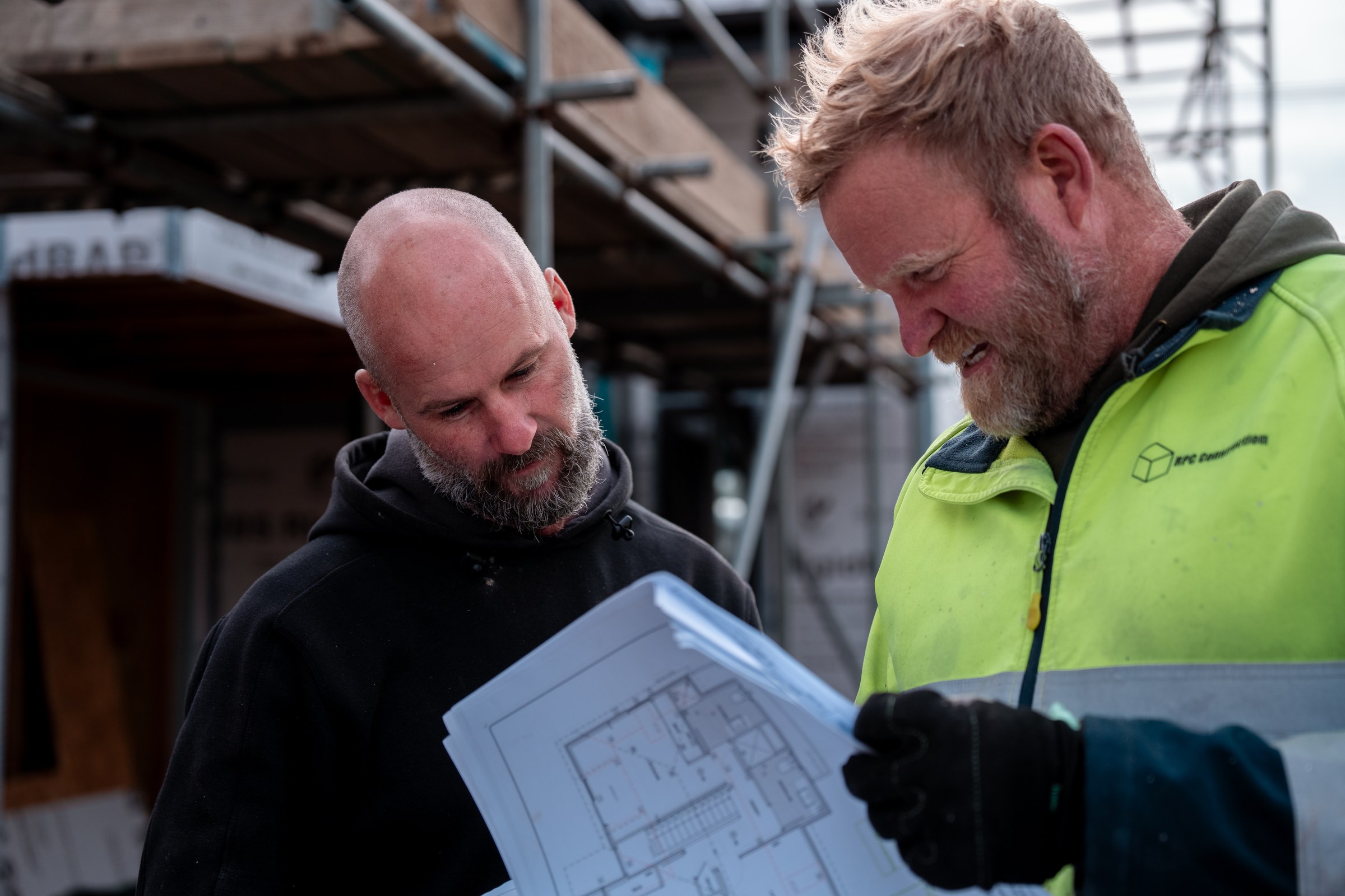 Two people looking at an open architectural floor plan held between them on a building site, one in an RPC Construction hi-vis jacket